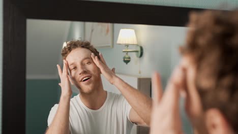 a handsome adult man standing in front of a mirror applies face cream with his hands