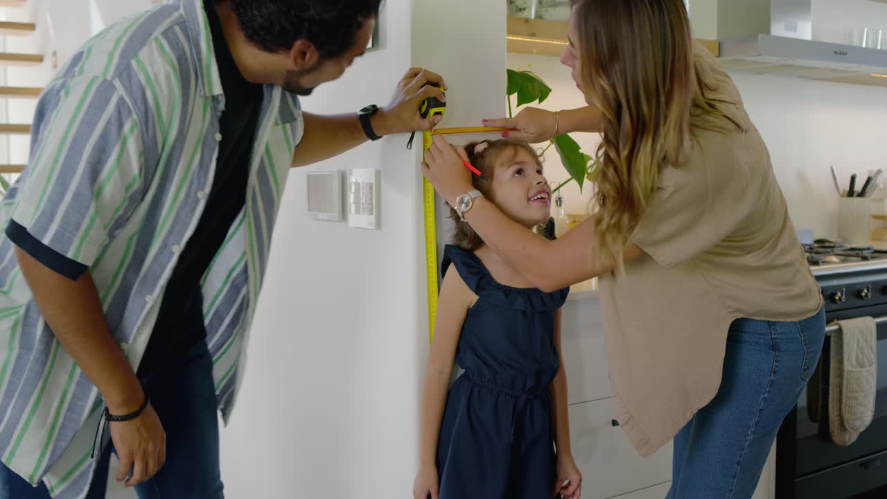 Diverse family measuring daughter in kitchen, dad with yellow tape, mom using pen to record height