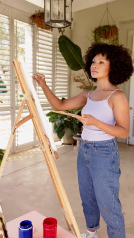 vertical video: painting on canvas, woman standing at easel in bright room with plants