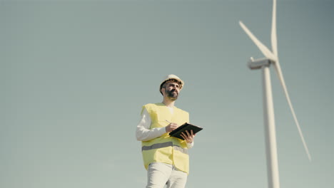 a young tech-savvy caucasian engineer in a reflective vest inspects wind turbines on a sunny day, showcasing the future of clean energy production