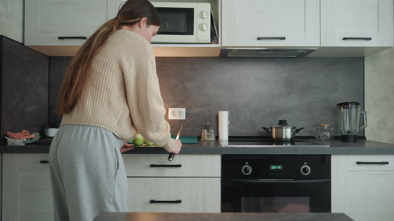 Donna bianca incinta che taglia le mele su un tagliere, in piedi in una cucina moderna, indossa un cardigan comodo e una lunga treccia, preparazione attiva per uno spuntino sano, mani concentrate e pancia visibile, bagliore caldo della lampada.