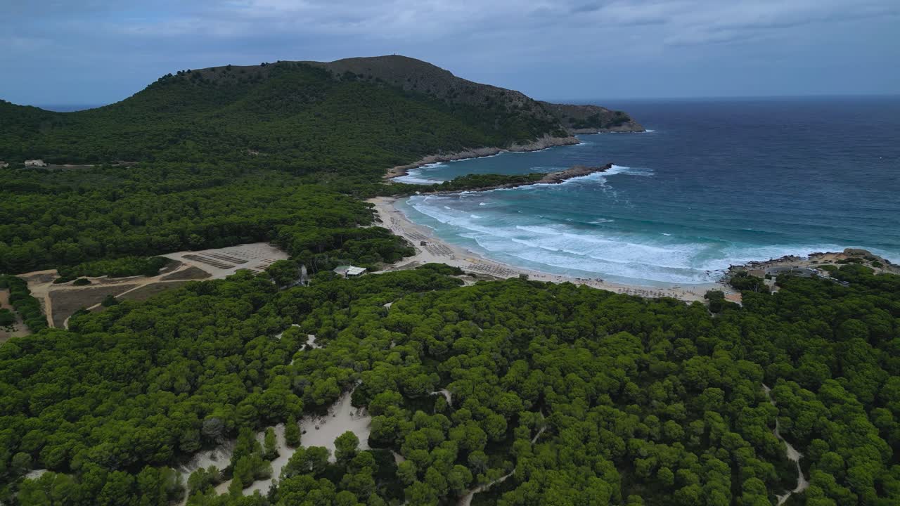 Cala Agulla, Maiorca. La scena mostra onde impetuose, il contrasto tra la lussureggiante pineta verde, le calette frastagliate e lo sviluppo del resort turistico sotto un cielo nuvoloso e drammatico