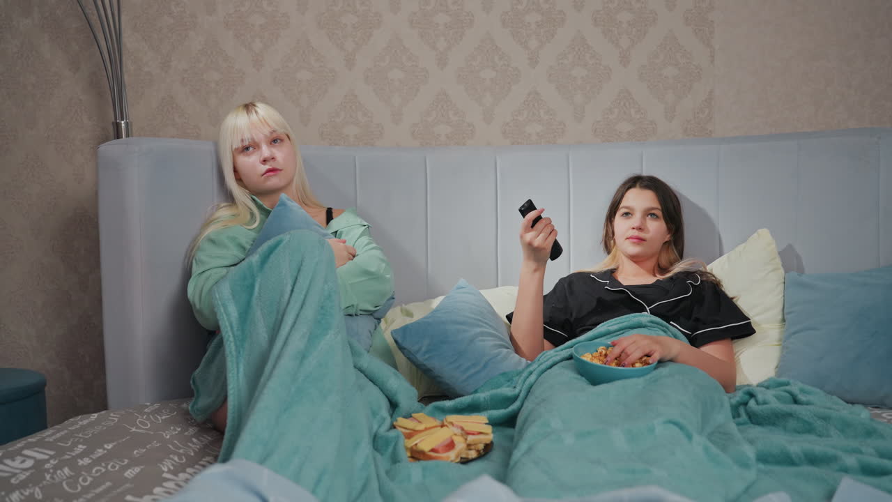 Two teenage girls in pajamas lying in bed watching TV with bored expressions, one holds remote control while other hugs pillow, popcorn bowl rests on blanket as they search for interesting