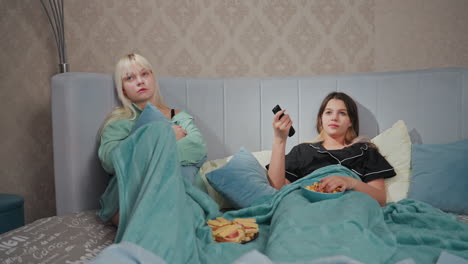 two teenage girls in pajamas lying in bed watching tv with bored expressions, one holds remote control while other hugs pillow, popcorn bowl rests on blanket as they search for interesting