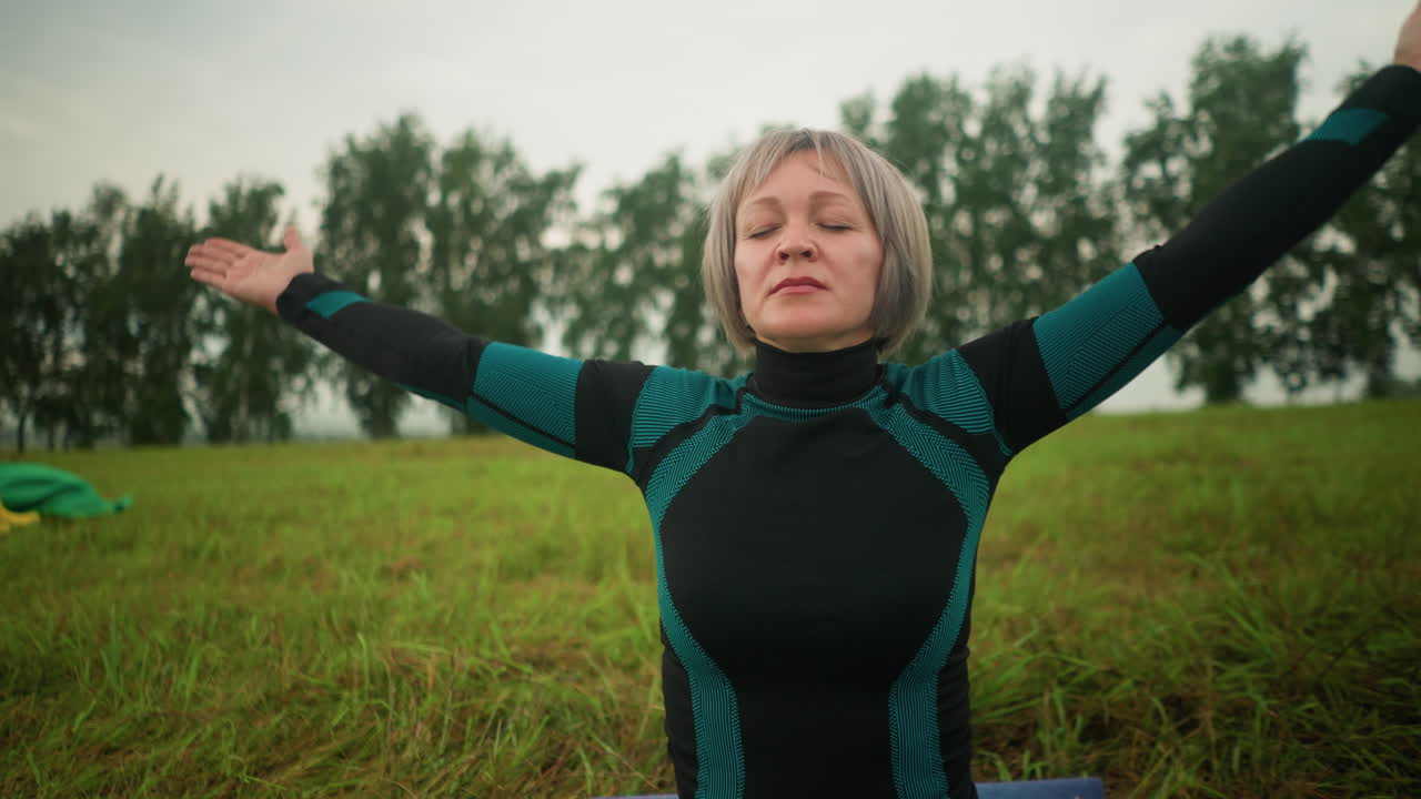 Close up of woman seated outdoors in grassy field, eyes closed, meditating with arms raised at shoulder level, practicing yoga under cloudy sky, yellow boots and green cloth visible in background