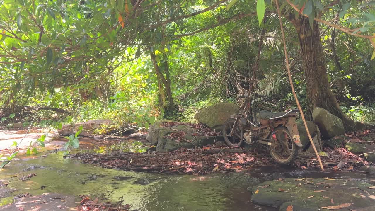 Old motorbike parked beside a rocky stream in dense tropical jungle