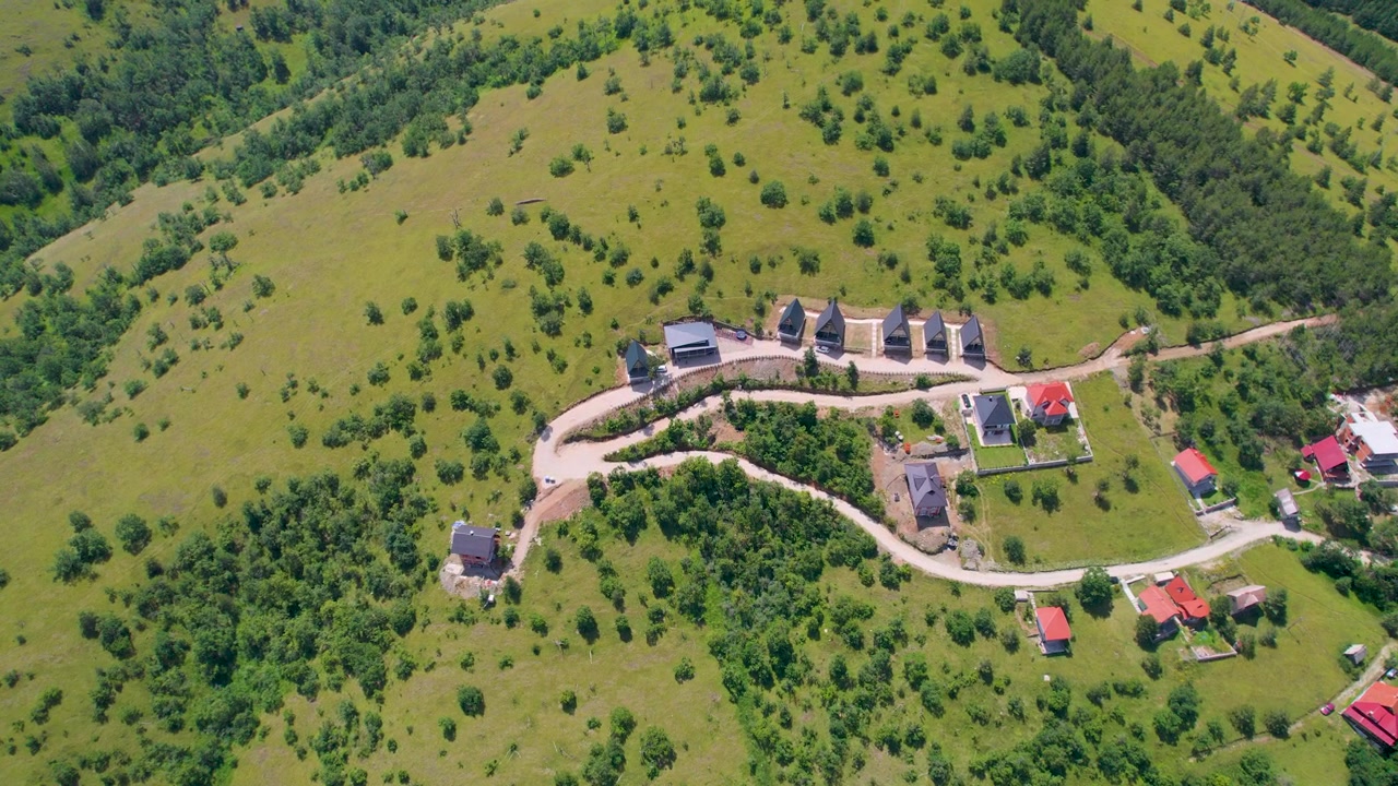 Et næsten lodret droneperspektiv cirkler et afsidesliggende bjergresort med klynger af hytter og en snoet adgangsvej midt i grønne bakker i Zlatibor, Serbien.