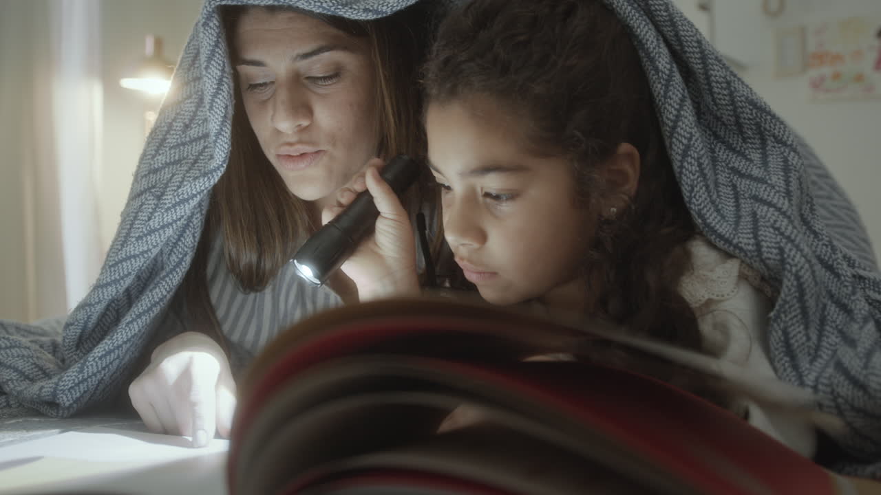 Little Girl Holding Flashlight as Mom Reading Fairytale in Bed at Night