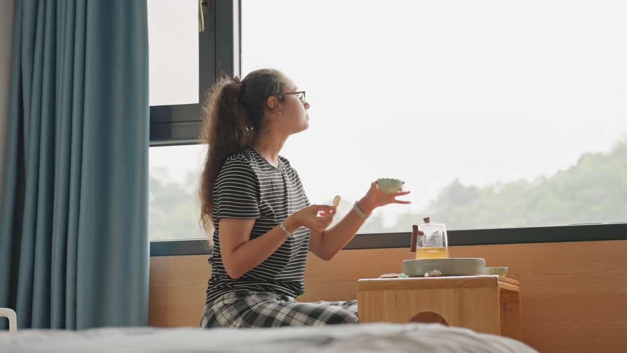 Tranquila habitación de hotel con vistas serenas a la naturaleza. Una mujer relajada bebe té mientras las colinas brillan suavemente en el exterior. Ambiente apacible de hotel con la mujer saboreando su desayuno mientras observa paisajes verdes.