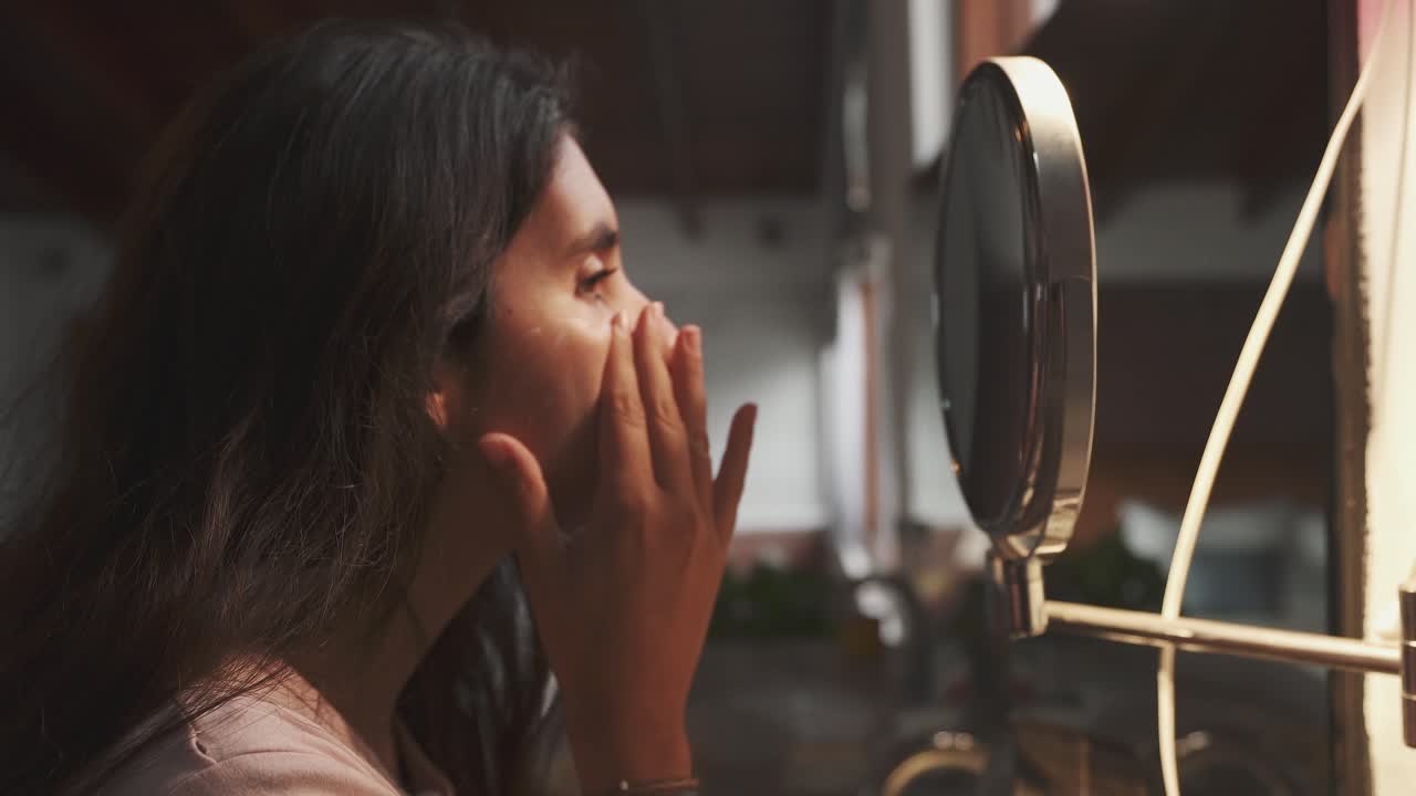 매력적인 브루네트 소녀는 욕실에 있는 둥근 거울을 보며 부드럽게 얼굴에 크림을 바르고 있다