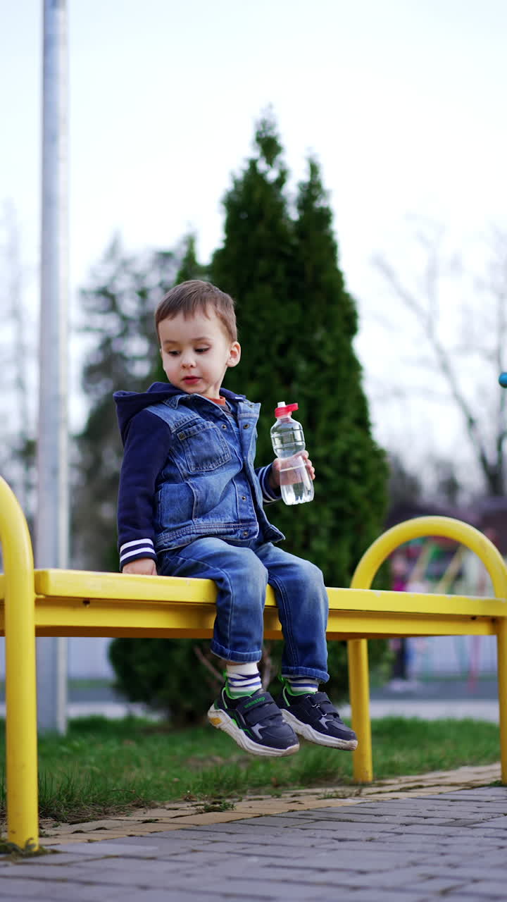 Two year old toddler boy in jeans clothes takes a bottle from the bench. Kid drinks water and talks. Vertical video.