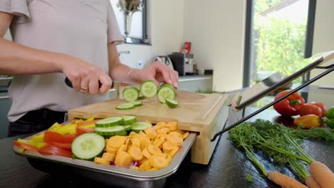 Toma-En-órbita-De-Una-Persona-Cortando-Pepinos-En-Una-Tabla-De-Cortar-Con-Otras-Verduras-Frescas-Preparadas,-En-Una-Cocina-Casera-Moderna-Y-Limpia-En-Un-Día-Soleado