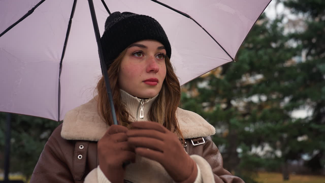 Female with eyes closed holding umbrella over head in cold weather, wearing black knit cap and brown shearling jacket, standing outdoors surrounded by green pine trees with soft light snowfall