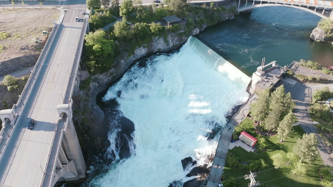 Cars cross a bridge above the dam in downtown Spokane. The waterfall drops sharply below, surrounded by trees and buildings