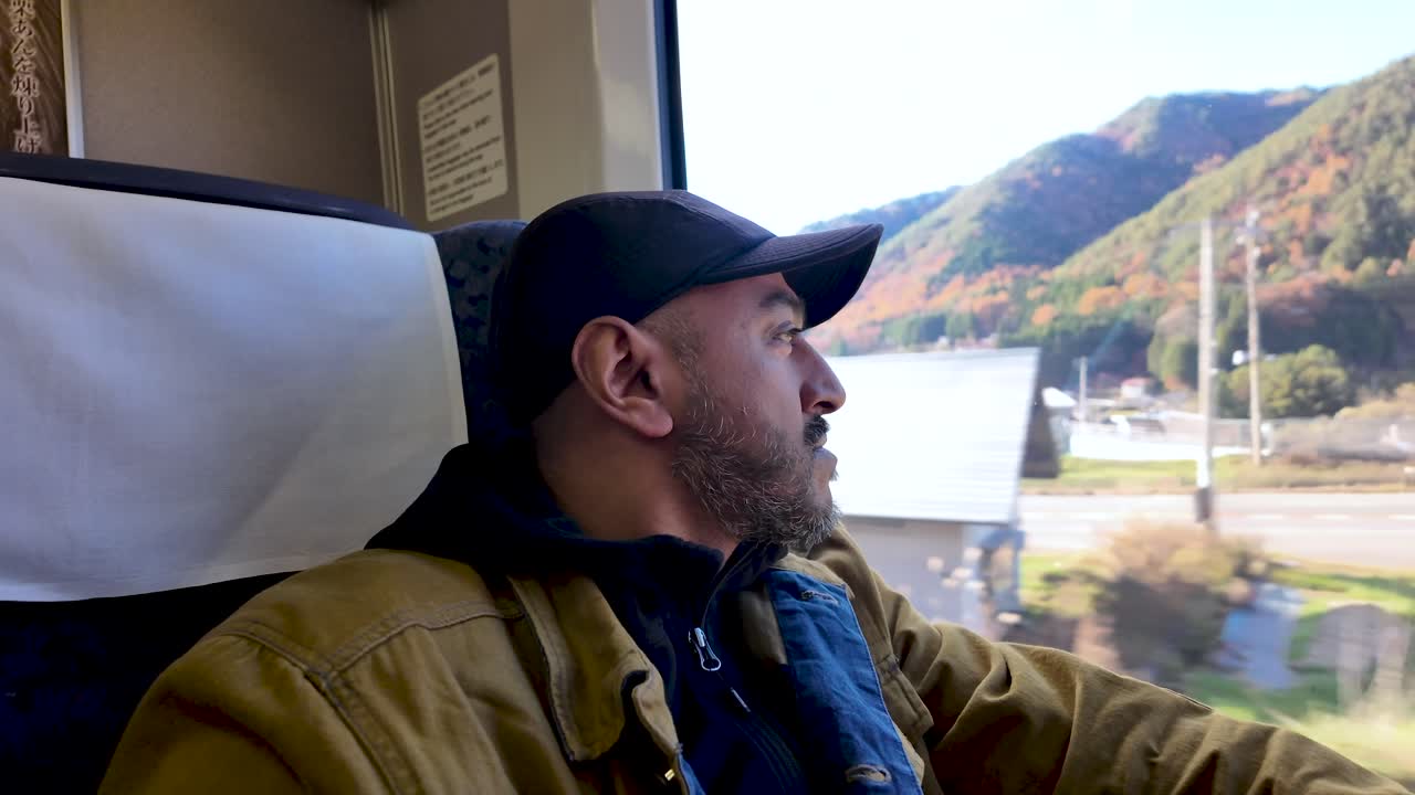 South Asian man gazes out train window at picturesque landscape during solo travel journey.