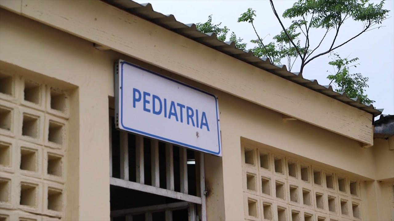Close-up em ângulo baixo de uma placa desgastada "PEDIATRIA" no exterior de um hospital em Moçambique, destacando a infraestrutura de saúde, a arquitetura da instalação e elementos do clima tropical.