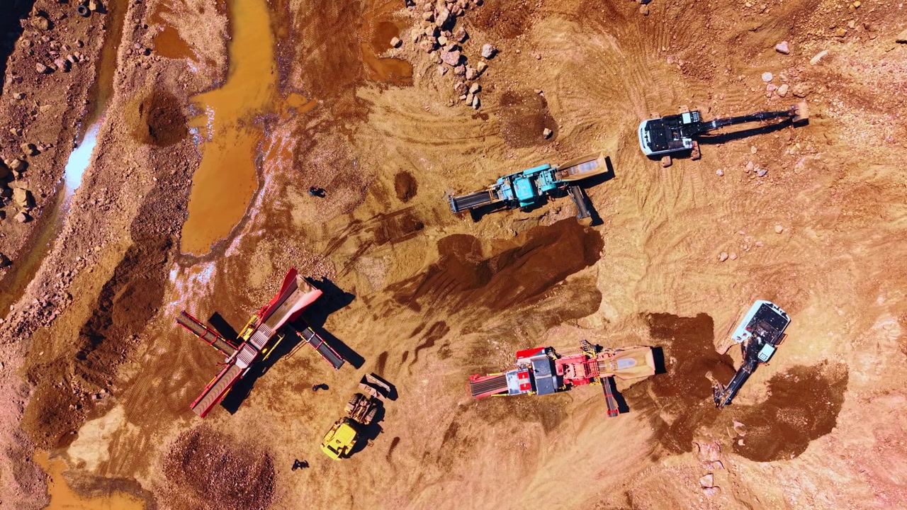 Mining equipment operating inside an open pit stone quarry. Vertical view of heavy excavators, mobile crushers, brown soil, rock piles and direct daylight.