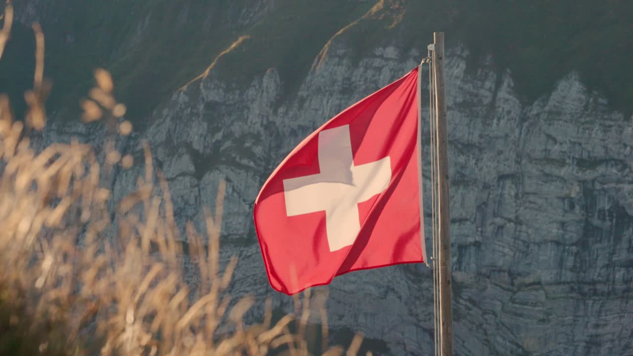 Rote Schweizer Flagge mit weißem Kreuz weht stolz am Fahnenmast vor dramatischen Kalksteinfelsen. Die Beleuchtung der goldenen Stunde erhellt zerklüftete Alpengipfel und Herbstgras im Vordergrund und schafft eine beeindruckende Kulisse.