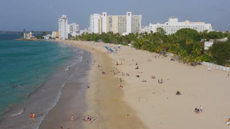 An-aerial-view-of-a-coastal-section-of-San-Juan,-Puerto-Rico-with-tourist-lounging-on-the-beach-and-resorts-in-the-background