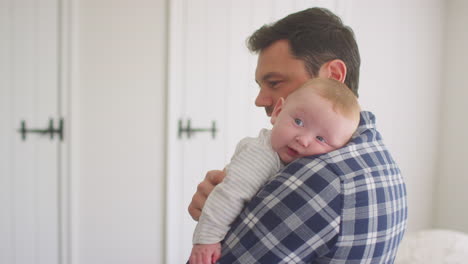 Loving-father-cuddling-smiling-baby-son-resting-over-shoulder-at-home-together---shot-in-low-motion