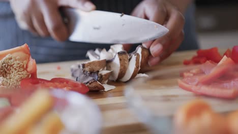 Midsection-of-african-american-woman-in-apron-chopping-mushrooms-in-kitchen,-slow-motion