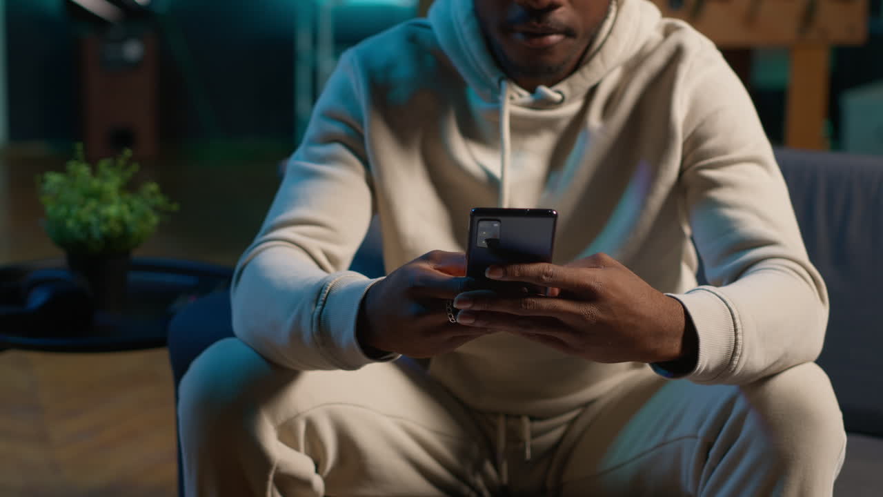 Relaxed black freelancer chatting with friends on his smartphone via texts, sitting on the couch. Young adult showcasing laughter and leisure, staying entertained while working remotely. Camera A.