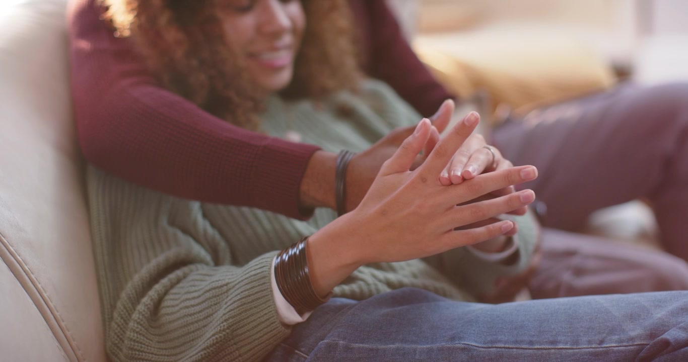 Couple sitting on sunlit sofa, man wrapping arm, woman moving hands, him touching fingers to bond