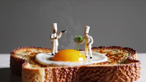 two miniature chefs stand on a fried egg over a slice of toast, one holding a serving tray and the other presenting a small plant, with steam rising from the scene, isolated on a white background