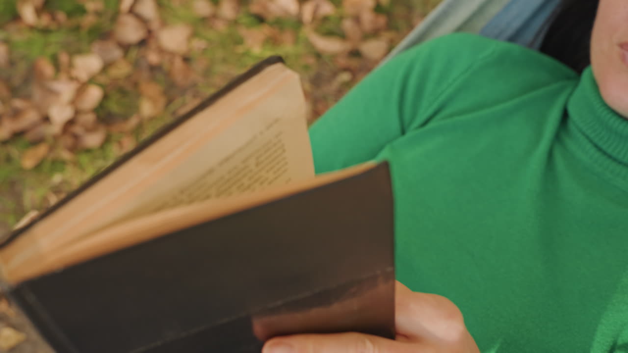 Donna in amaca che legge un libro, in un campeggio tranquillo con foglie cadute. Maglione verde, pagine che si girano lentamente. Primi piani del dorso del libro e delle mani, luce solare soffusa. Un tranquillo svago solitario per scrittori o lettori.