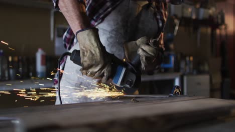 Midsection-of-caucasian-male-knife-maker-in-workshop-wearing-glasses-and-using-angle-grinder