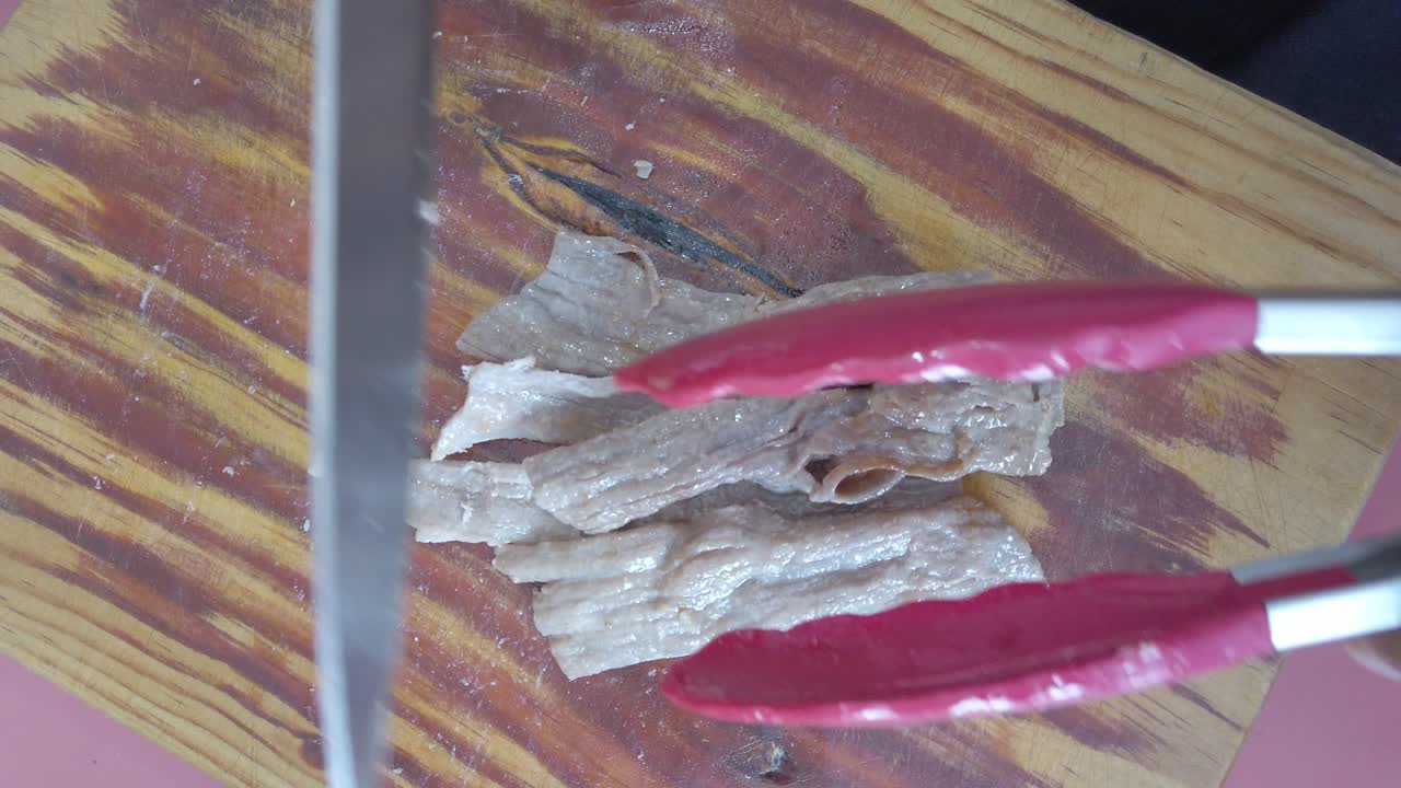 Slicing cooked meat on wooden board with kitchen tongs and knife