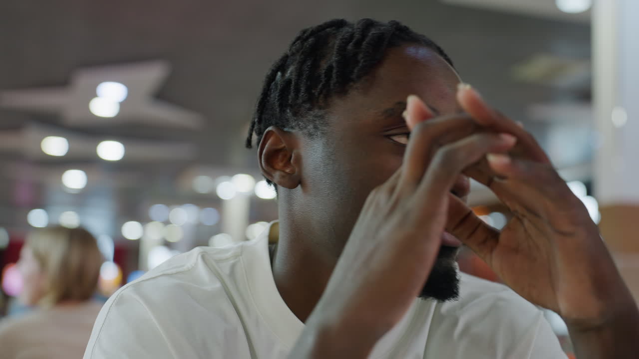 Young african man wearing white shirt closes eyes and tilts neck in playful exaggerated motion while seated inside bright modern indoor cafe with glowing ceiling lights and blurred background crowd