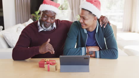 happy african american father and adult son in christmas hats having tablet video call, slow motion