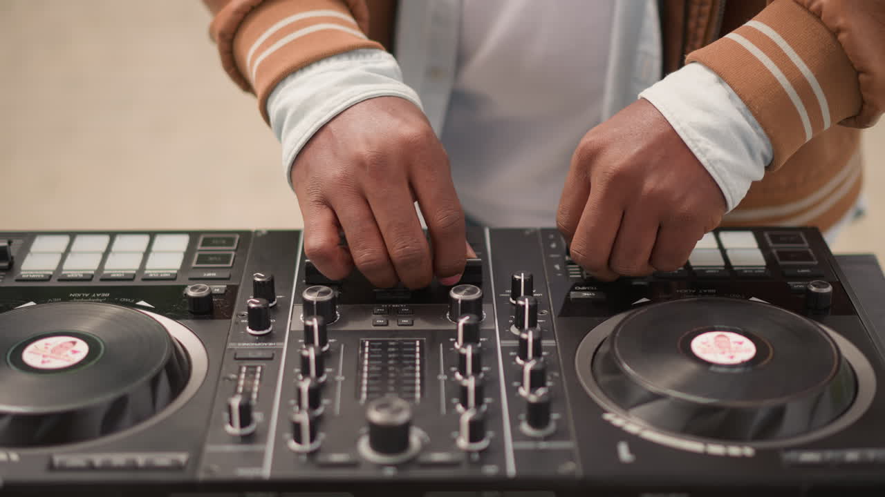 Close-up tangan DJ hitam sedang mixing kontroler di studio latihan yang intim, jaket vintage dan gelang, penyesuaian fader dan kenop yang presisi, jog wheel vinil, cahaya ambient hangat, teknik terfokus.