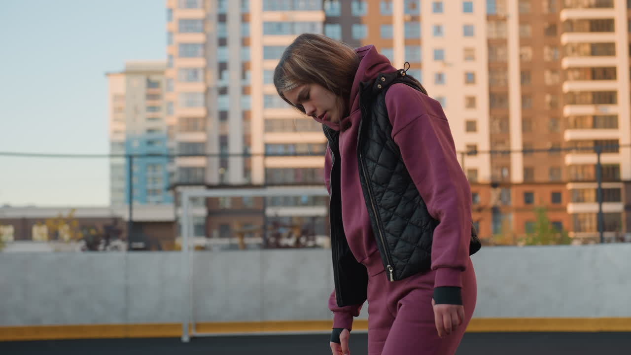 Medium shot of hoodie wearing female athlete looking down in purple tracksuit and black vest on outdoor round court with blurred city high rise buildings and fence at dusk under golden sunset light
