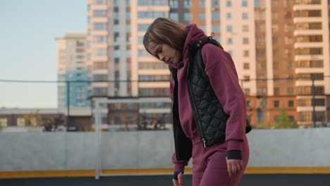 medium shot of hoodie wearing female athlete looking down in purple tracksuit and black vest on outdoor round court with blurred city high rise buildings and fence at dusk under golden sunset light
