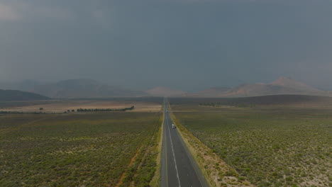 Los-Reenvíos-Vuelan-Por-Encima-De-La-Carretera-Que-Conduce-Directamente-En-Un-Paisaje-Llano-Con-Montañas-A-Lo-Lejos.-Poco-Tráfico-En-La-Carretera.-Sudáfrica