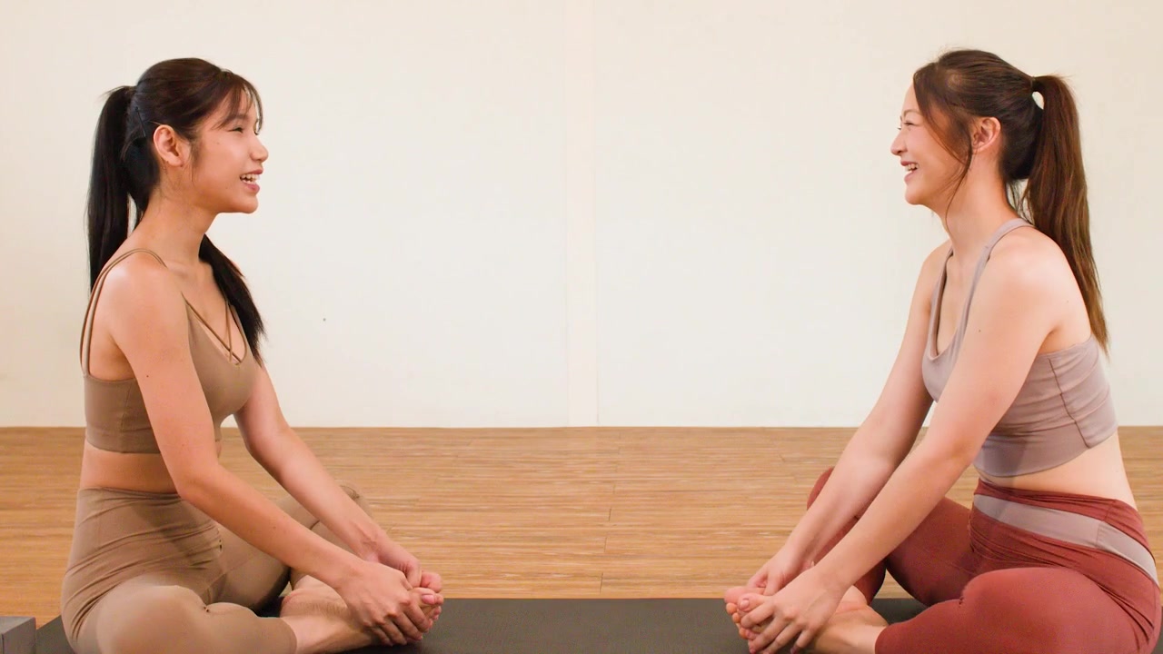 Due donne in abbigliamento sportivo eseguono allungamenti a farfalla su tappetini in uno studio yoga luminoso e minimalista.