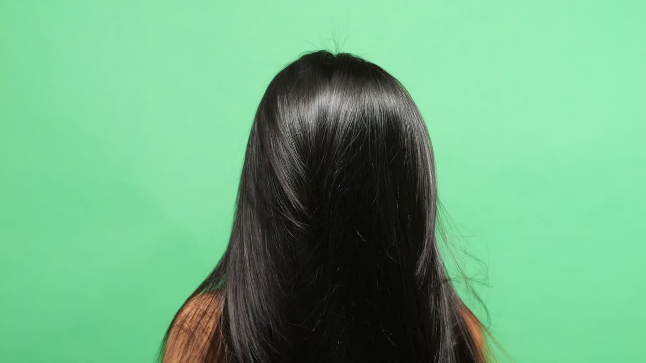 녹색 스크린 배경 스튜디오에서 그녀의 길고 검은 직선 건강한 머리카락을 뒤집는 여자의 가까운 뒷면