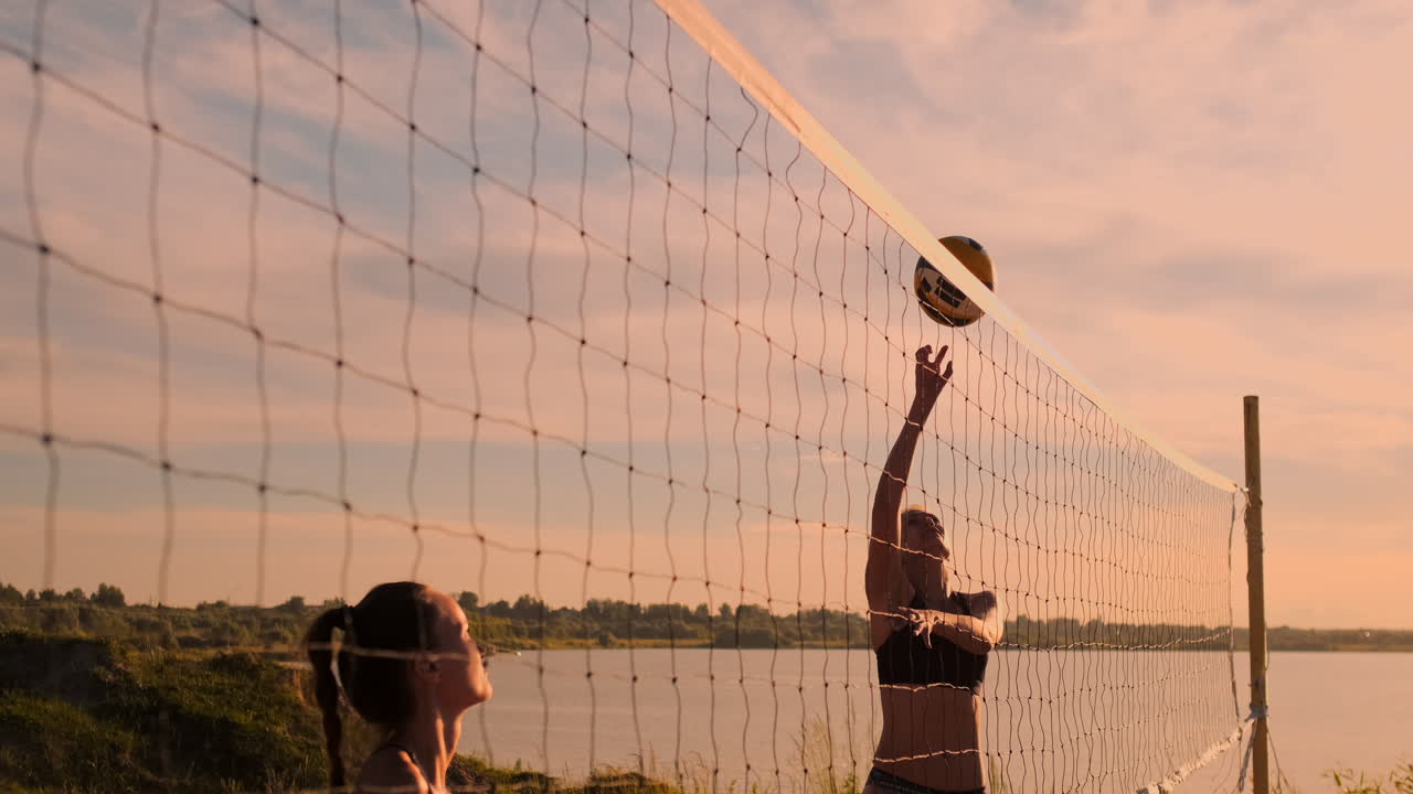 느린 움직임 낮은 각도 근접 태양 플레어: 해변 볼리볼을하는 운동 소녀가 공중에 뛰어들고 아름다운 여름 저녁에 네트 위에 공을 때립니다.