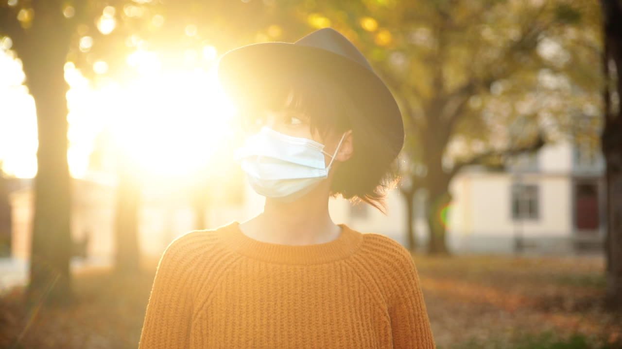 Young brunette woman walking under trees in autumn with sun light, wearing a mask