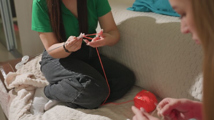 caucasian friend teaching knitting on couch in bedroom setting, green shirt instructor demonstrating stitches with red yarn ball, hands closeup, soft blanket, warm natural light, relaxed tutorial