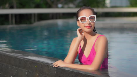 a healthy young woman in a pink one-piece swimsuit, lounges along the edge of an infinity pool