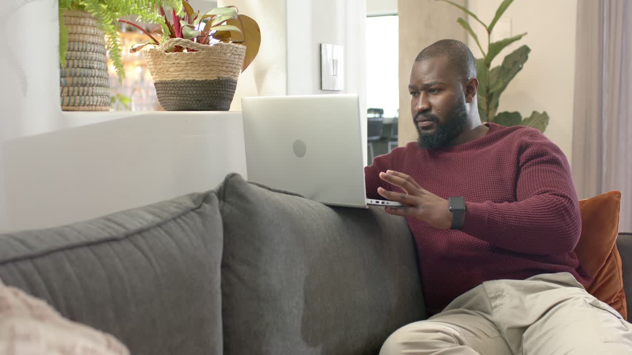Mid adult African American man placing laptop on sofa, typing for work at home, copy space