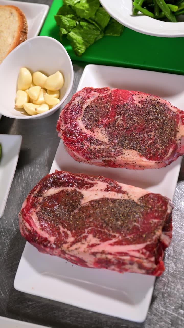 Two raw ribeye steaks with seasoning on a plate. Top view of ribeye steaks with pepper and seasoning placed on a white plate
