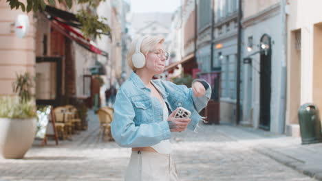 overjoyed blonde woman in wireless headphones choosing listening music holding smartphone on street