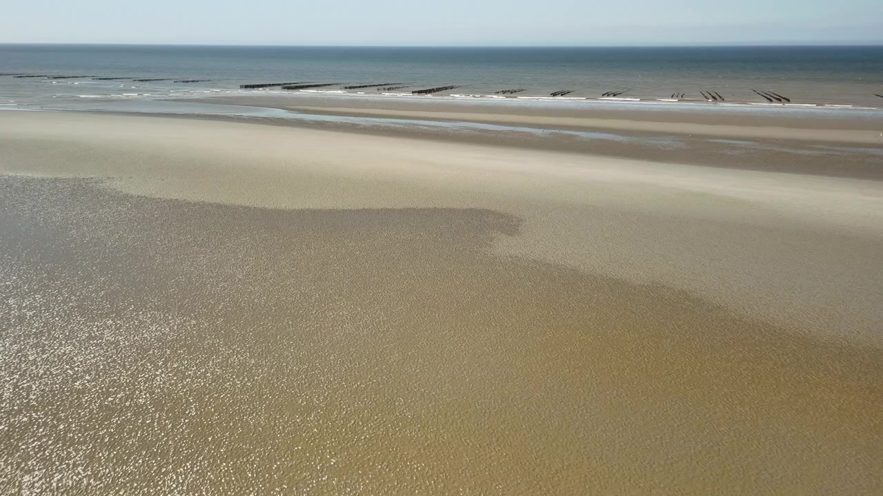 Lav droneflyvning over sandede vadeflader mod havet med en blid panorering mod højre, der afslører traditionelle muslingefarme-pæle nær Saint-Quentin-en-Tourmont, Picardiet, Frankrig.