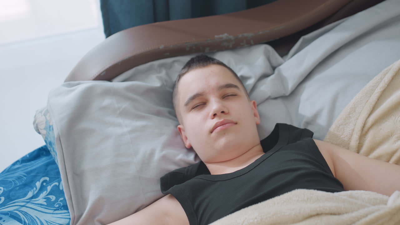 Kid lying in bed stretching under beige blanket with eyes closed showing peaceful rest and relaxation in cozy bedroom with soft natural light creating calm morning atmosphere of comfort