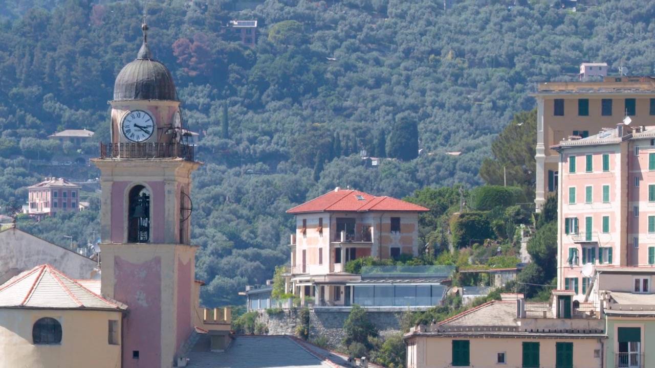 La luce del sole illumina colorati edifici mediterranei e una storica torre dell'orologio su un ripido pendio.