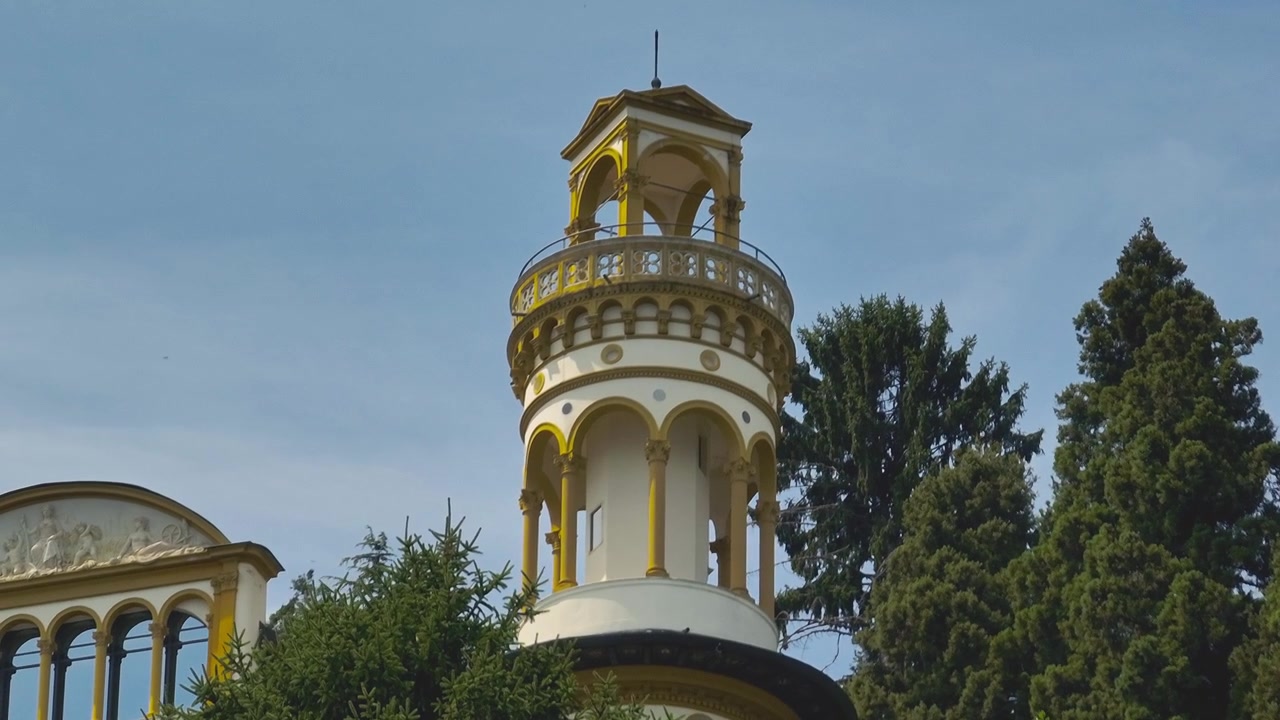 Vista aérea aproximada de drone de uma elegante torre de vila italiana exibindo detalhes arquitetônicos arredondados contra um céu azul claro enquanto pássaros voam, destacando a arquitetura europeia atemporal.